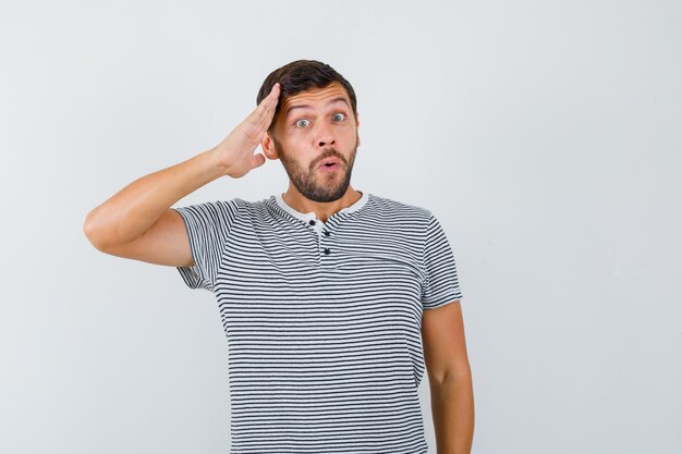 Jovem em t-shirt listrada, mostrando o gesto de saudação e olhando maravilhado, vista frontal.