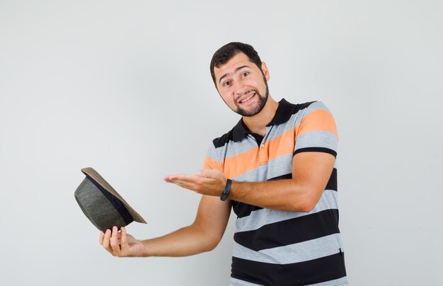 Jovem em t-shirt, mostrando seu chapéu e olhando alegre, vista frontal.