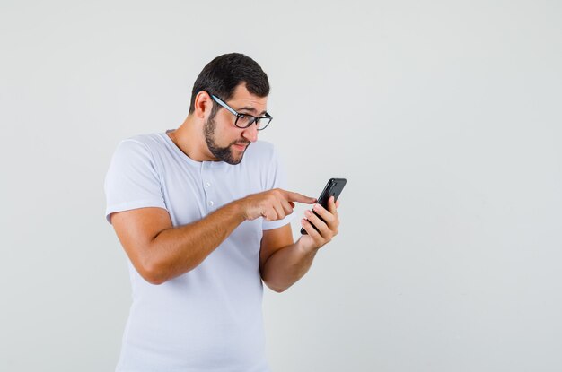 Jovem em t-shirt, óculos, olhando para o telefone e olhando com foco, vista frontal.