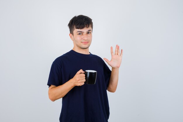 Jovem em t-shirt preta segurando uma xícara de chá, mostrando a palma da mão e olhando confiante, vista frontal.