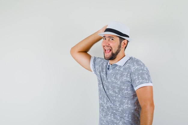 Jovem em t-shirt segurando a mão em seu chapéu e olhando alegre, vista frontal.