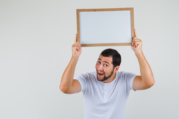 Foto grátis jovem em t-shirt segurando um quadro branco sobre a cabeça e olhando alegre, vista frontal.