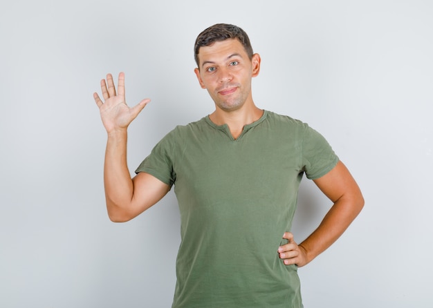 Foto grátis jovem em t-shirt verde exército, levantando a palma da mão em gesto de olá e olhando positivo.