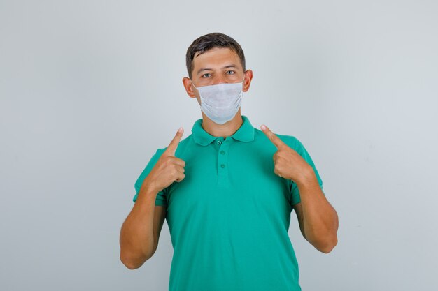 Jovem em t-shirt verde, mostrando sua máscara médica e olhando cuidadosa, vista frontal.