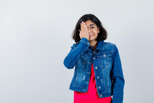 Jovem em t-shirt vermelha e jaqueta jeans cobrindo os olhos com a mão e parecendo feliz, vista frontal.