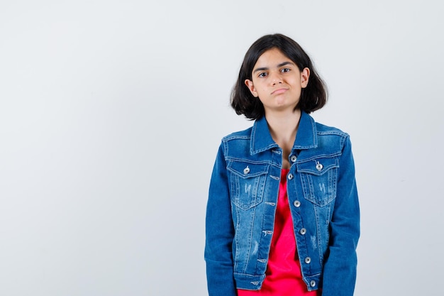 Jovem em t-shirt vermelha e jaqueta jeans em pé em linha reta e posando para a câmera e olhando feliz, vista frontal.