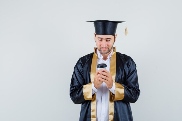 Jovem em uniforme de pós-graduação, olhando para o copo de café de papel, vista frontal.