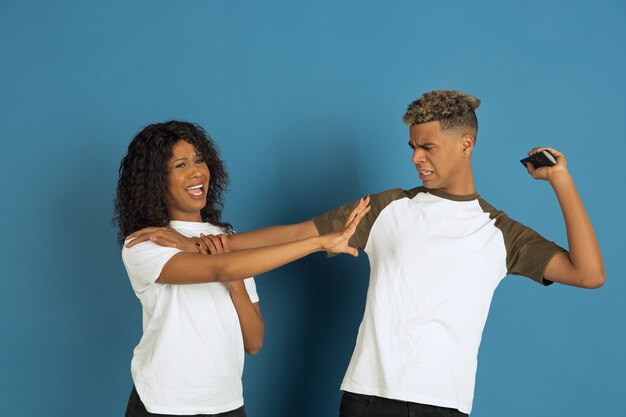 Jovem emocional afro-americano e mulher em roupas casuais brancas, posando sobre fundo azul. Casal bonito. Conceito de emoções humanas, expressão facial, relações, anúncio. Assistam TV juntos.
