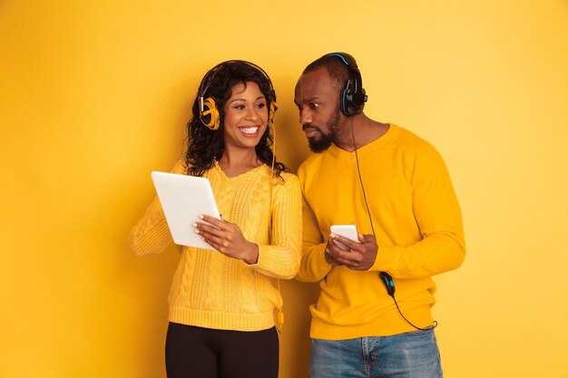 Jovem emocional afro-americano e mulher em roupas casuais brilhantes sobre fundo amarelo. Casal bonito. Conceito de emoções humanas, expressão facial, relações, anúncio. Usando tablet e smartphone.