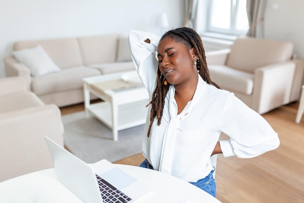 Jovem empresária africana tendo dor nas costas enquanto está sentado na mesa de escritório Empresária segurando suas costas enquanto trabalhava no laptop na mesa de escritório