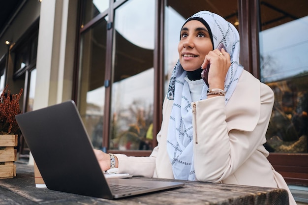 Jovem empresária árabe atraente em hijab falando no smartphone durante o trabalho no laptop na rua da cidade