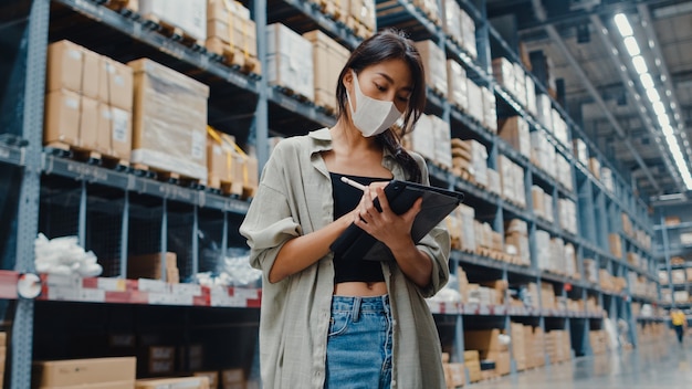 Jovem empresária asiática usando máscara facial no armazém usando tablet digital, verificando o inventário