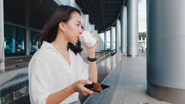 Jovem empresária bem-sucedida da Ásia em roupas de escritório da moda, segurando um copo de papel descartável com uma bebida quente e usando um telefone inteligente enquanto está ao ar livre na cidade moderna urbana