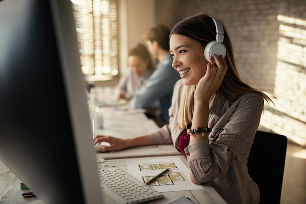 Jovem empresária feliz trabalhando em um computador e ouvindo música em fones de ouvido no escritório seus colegas estão trabalhando em segundo plano