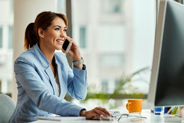 Jovem empresária feliz trabalhando no PC desktop e se comunicando por telefone celular no escritório