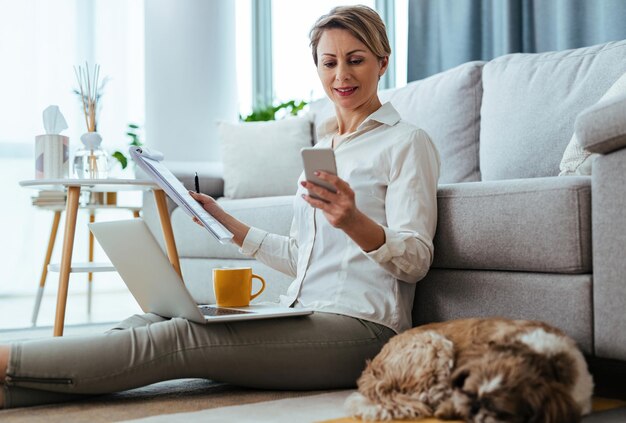 Jovem empresária mandando mensagens no smartphone enquanto está sentada no chão e trabalhando em casa Seu cachorro está dormindo ao lado dela