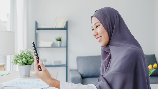 Jovem empresária muçulmana da Ásia usando um telefone inteligente, fala com um amigo por videochat, brainstorm, reunião on-line, enquanto trabalha remotamente em casa na sala de estar.