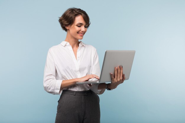 Jovem empresária muito sorridente com cabelo curto escuro na camisa branca trabalhando no laptop sobre fundo azul isolado