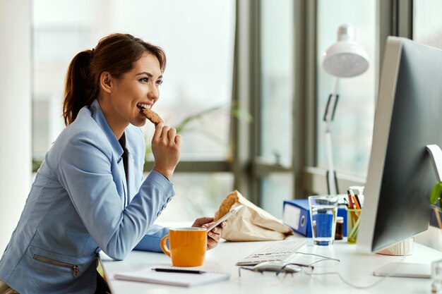 Jovem empresária usando telefone celular enquanto come um biscoito em sua mesa de escritório