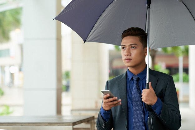 Jovem empresário asiático com guarda-chuva em pé na rua com o smartphone e desviar o olhar