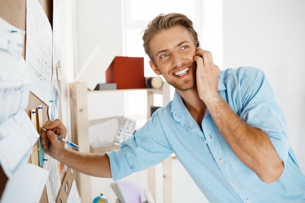 Jovem empresário bonitão, escrevendo no jornal fixado ao quadro de cortiça, falando no telefone, sorrindo.