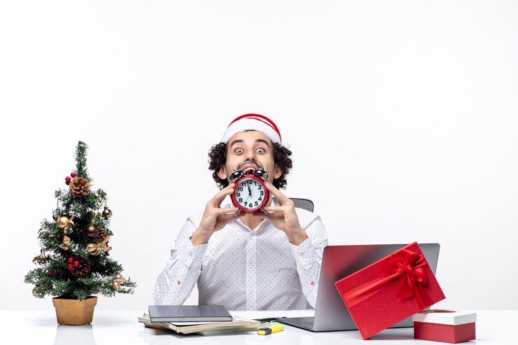 Foto grátis jovem empresário com chapéu de papai noel e mostrando o relógio olhando algo com cuidado e sentado no escritório sobre fundo branco