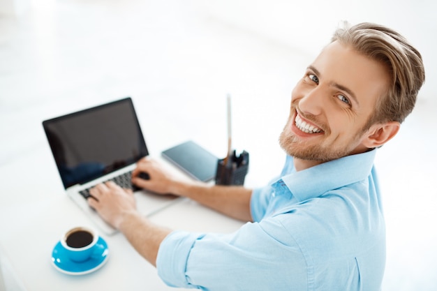 Jovem empresário confiante alegre bonito sentado à mesa, trabalhando no laptop com xícara de café de lado. sorridente. Interior de escritório moderno branco