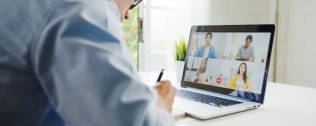 Jovem empresário da Ásia usando laptop fala com colegas sobre o plano de uma reunião de videochamada enquanto trabalha em casa na sala de estar. Auto-isolamento, distanciamento social, quarentena para prevenção do vírus corona.