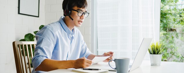 Jovem empresário da Ásia usando laptop fala com colegas sobre o plano de videochamada enquanto trabalha em casa na sala de estar. Auto-isolamento, distanciamento social, quarentena para prevenção do vírus corona.