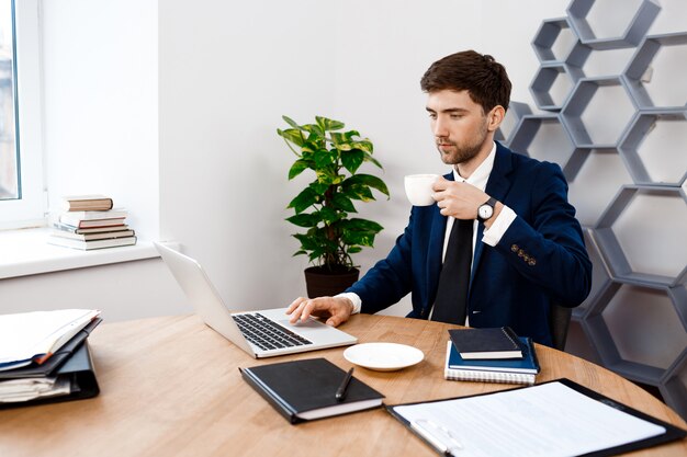 Jovem empresário de sucesso sentado no laptop, fundo de escritório.