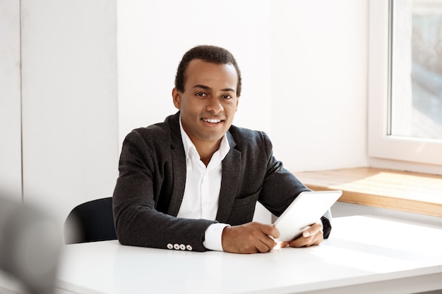 Foto grátis jovem empresário de sucesso sorrindo, segurando o tablet, sentado no local de trabalho