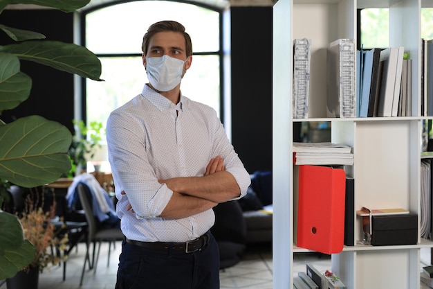 Jovem empresário em pé de máscara médica no escritório moderno.