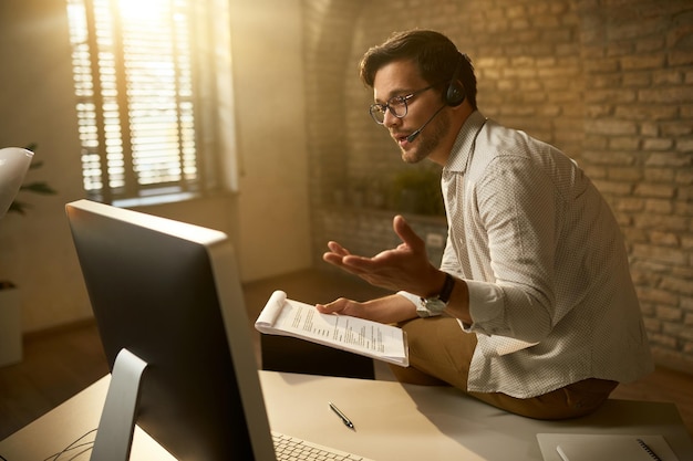 Jovem empresário falando sobre relatórios de negócios enquanto faz videoconferência no pc de mesa no escritório