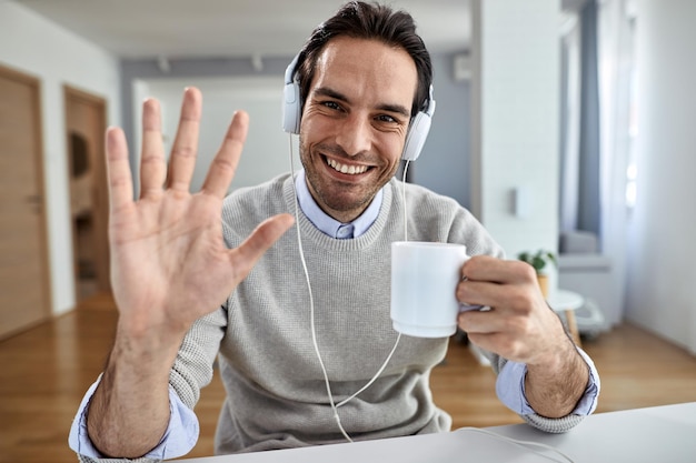 Jovem empresário feliz com fones de ouvido acenando para a câmera enquanto bebe café em casa