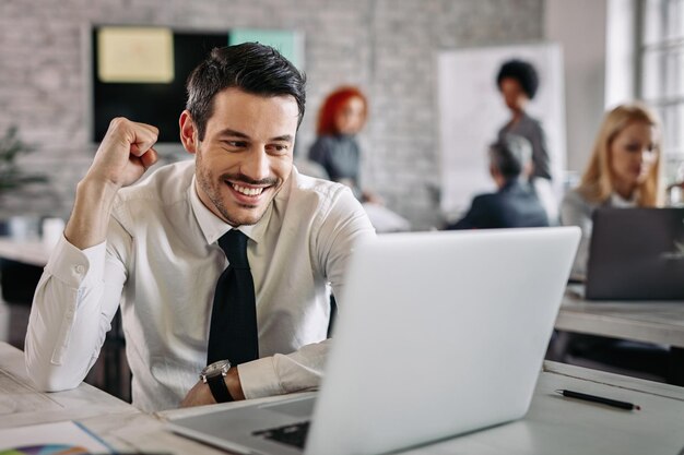 Jovem empresário feliz usando computador no escritório e comemorando boas notícias Há pessoas em segundo plano