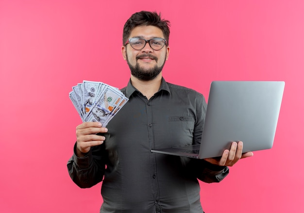 Foto grátis jovem empresário satisfeito usando óculos, segurando o laptop e o dinheiro isolado no fundo rosa