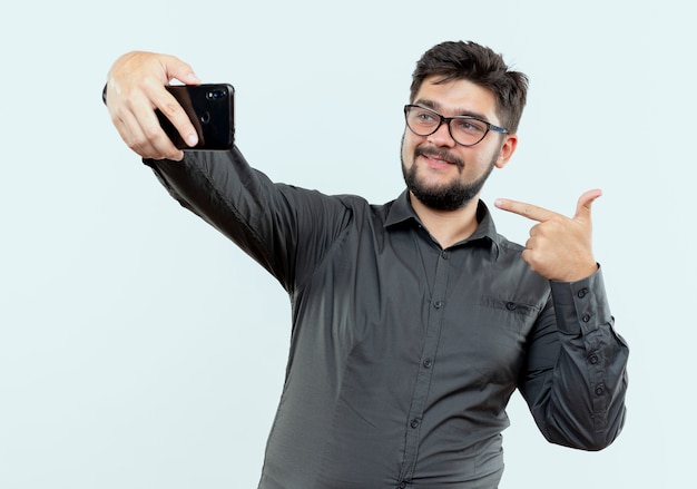 Jovem empresário satisfeito, usando óculos, tira uma selfie e aponta para si mesmo, isolado no fundo branco