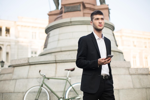 Jovem empresário sério em terno preto clássico e camisa branca com fones de ouvido sem fio segurando o celular na mão enquanto pensativamente olhando de lado com bicicleta retrô em fundo difuso ao ar livre