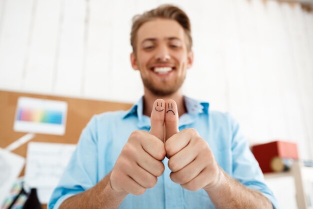 Jovem empresário sorridente bonito mostrando os polegares com desenhos de caretas. Concentre-se nas mãos. Interior de escritório moderno branco