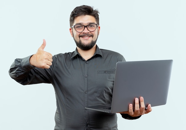 Jovem empresário sorridente de óculos, segurando o laptop com o polegar isolado no branco