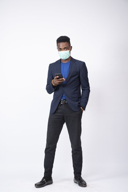 Jovem empresário vestindo terno e máscara facial usando seu telefone em frente a uma parede branca