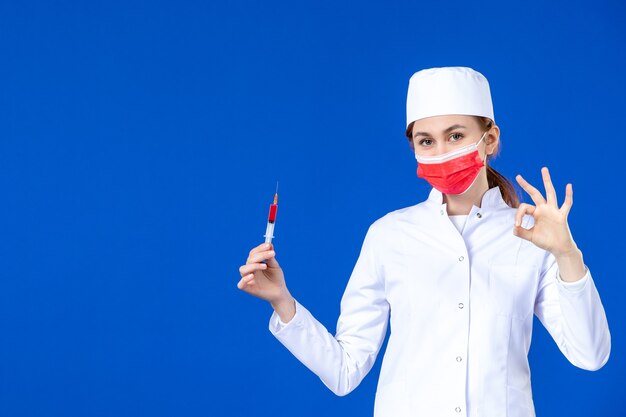 Jovem enfermeira de vista frontal em um terno médico branco com máscara vermelha e injeção nas mãos na parede azul