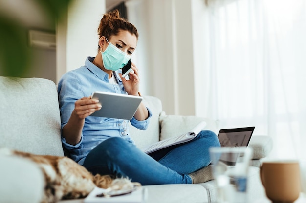 Jovem engenheira com máscara facial se comunicando no celular enquanto trabalhava no tablet digital em sua casa durante a epidemia de vírus