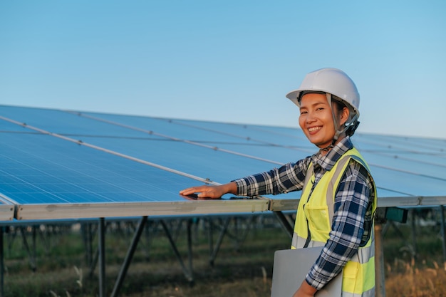 Jovem engenheira inspetora asiática tocando para verificar o painel solar fotovoltaico de operação na estação ao ar livre segurando o computador portátil e olhando a câmera com espaço de cópia de sorriso