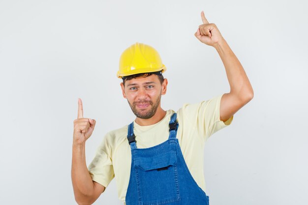 Jovem engenheiro apontando para cima e sorrindo de uniforme, vista frontal.