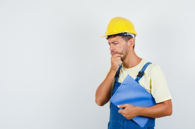 Jovem engenheiro de uniforme, segurando a prancheta e pensando, vista frontal.