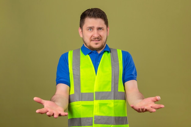 Foto grátis jovem engenheiro usando colete de construção parecendo confuso em pé com os braços e as mãos levantadas sobre um fundo verde isolado