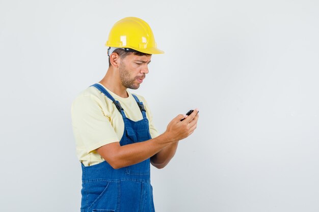 Jovem engenheiro usando telefone celular de uniforme e parecendo preocupado, vista frontal.