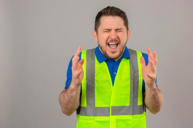 Jovem engenheiro vestindo colete de construção, louco e louco, gritando e gritando com expressão agressiva e braços levantados sobre um fundo branco isolado