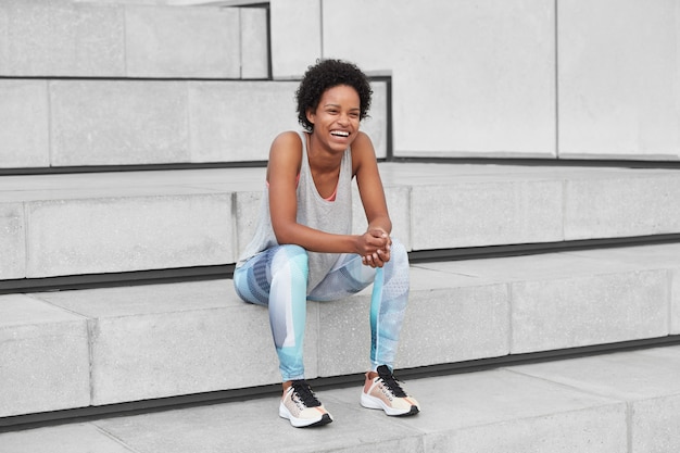 Jovem engraçado alegre com pele morena saudável, cabelo encaracolado, vestido com roupas esportivas, tênis, ri de algo positivo, conversa com um amigo após o treino de verão ao ar livre, senta na escada